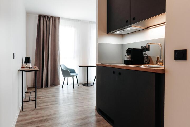 Minimalist interior of an AC Collection apartment at Appart'City, featuring a modern kitchenette with sleek black cabinetry, a coffee machine, a desk with a blue chair, and brown curtains, conveying a clean and functional ambiance.