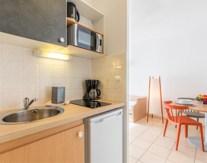 Fully equipped kitchen in an AC Classic aparthotel by Appart'City, featuring light wood cabinets, a built-in microwave, coffee maker, stainless steel sink, and an under-counter refrigerator, adjacent to a dining area with a round table and orange chairs on tiled flooring.