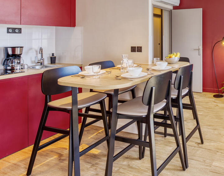 Dining area in an AC Confort apartment with a tall wooden table, high black chairs, breakfast plates and cups, a fully equipped kitchen with red cabinets in the background, providing a warm setting for meals.