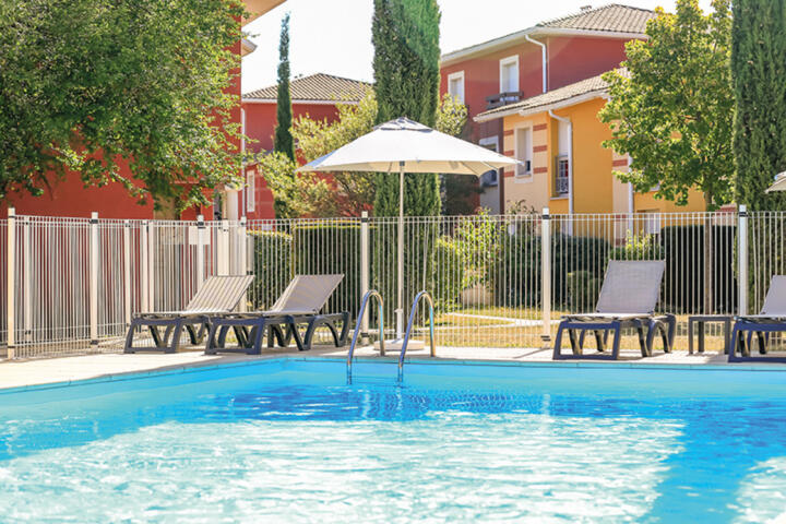 Sunny outdoor pool at Appart'City, surrounded by a safety fence, with comfortable loungers under a white umbrella, providing a relaxing area for families staying.