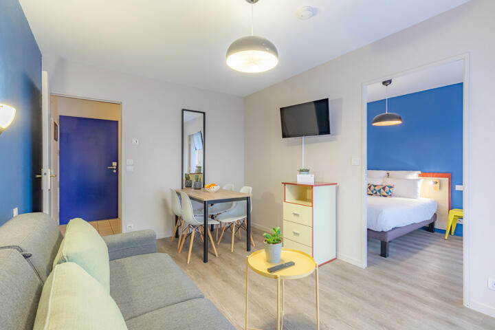 Modern and colorful interior of an Appart'City apartment designed for family stays, featuring a living area with a gray sofa, a yellow coffee table, a dining area with four chairs and a table, and a separate bedroom with a double bed in the background.